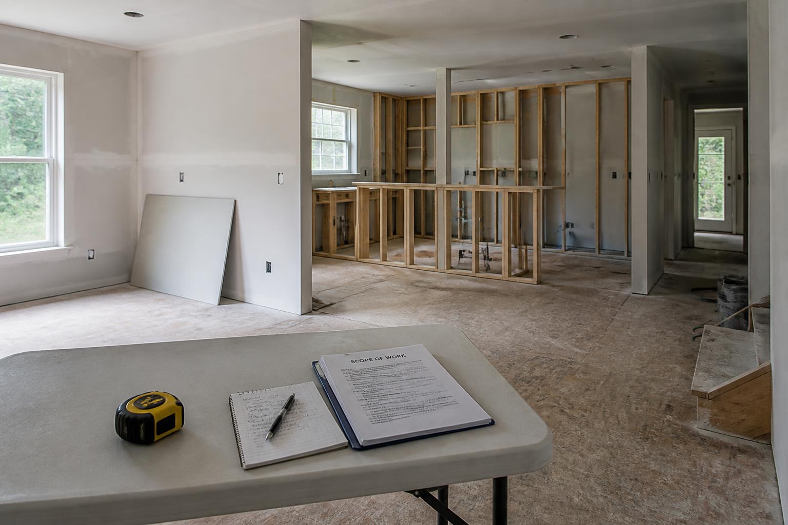 Interior of a house mid-renovation with framing, bare subfloor, and scope-of-work paperwork